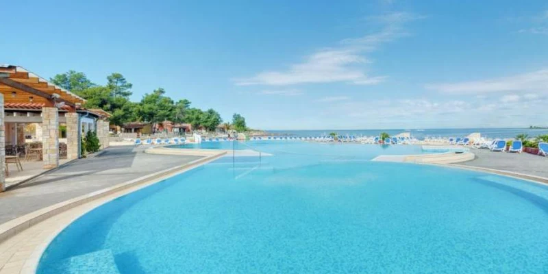 A large, curved swimming pool with surrounding lounge chairs overlooks a scenic lake; adjacent is a shaded restaurant area at Stella Plava Laguna