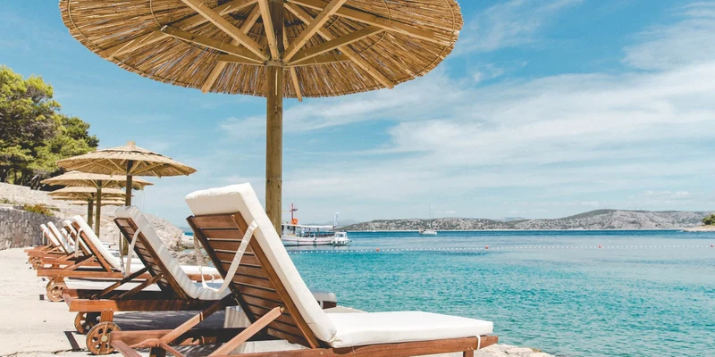 Wooden loungers resting under straw parasols near a serene ocean with distant boats and hills at Obonjan Island Resort