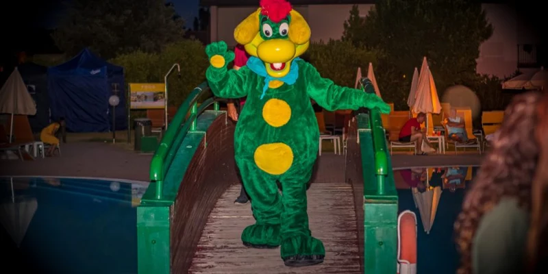 A green dragon mascot walks on a bridge near poolside loungers and umbrellas at Residence Eden