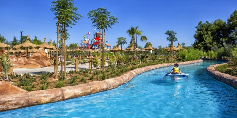 A person floats on a raft in a winding blue water river, surrounded by trees and tropical-themed landscaping at Amadria Park Camping Sibenik