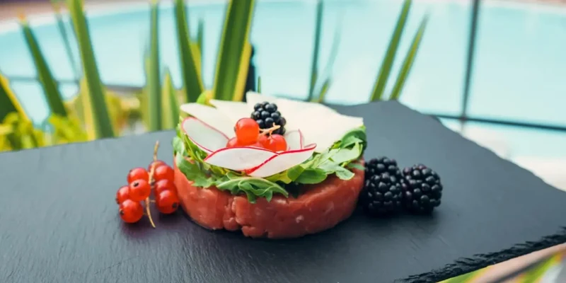 Elegant tartare garnished with radishes, red currants, and blackberries on slate plate, poolside greenery in background at Vacanze Glamping Boutique