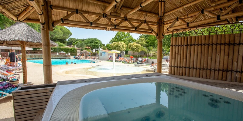 A jacuzzi under a bamboo hut overlooking a pool area with people sunbathing and swimming at Parc Mogador