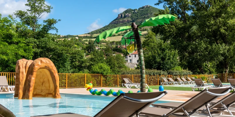 Pool loungers surround a splash pad with rock feature and palm tree fountain; mountains and greenery in the background at Le Millau Plage