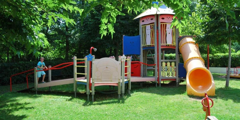 A child climbs a ramp towards a colorful playground structure surrounded by lush green trees at Residence Sognu di Rena