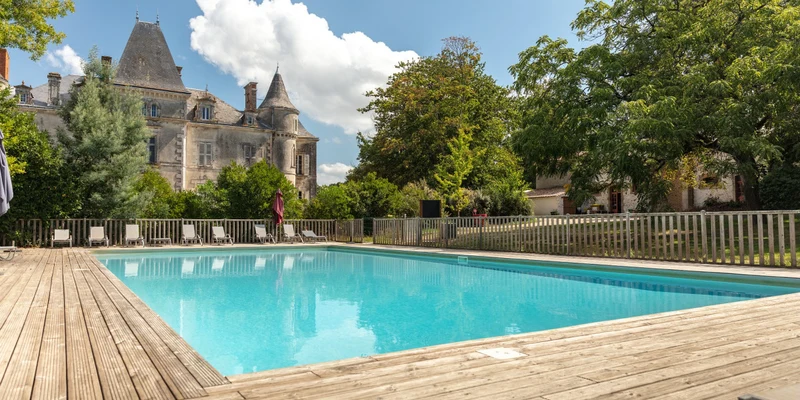 A serene swimming pool surrounded by wooden decking and lounge chairs, set near a large, historic chateau at Marvilla Parks Château la Forêt