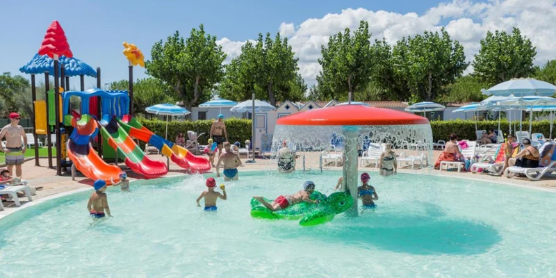 Children play in a shallow pool with colorful slides; adults relax on sun loungers under umbrellas at Riva Nuova