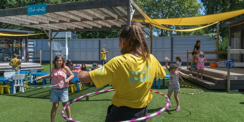 Instructor hula-hooping with children in a grassy, outdoor play area, under a wooden canopy with surrounding picnic tables at Les Mathes