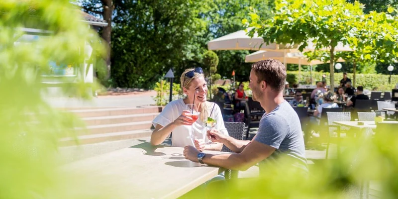 Two people enjoy drinks at an outdoor café, surrounded by trees and other patrons at Landal Sonnenberg