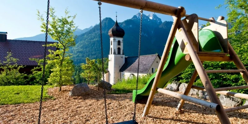 Children's playground with swings and slide on wood chips, set against mountain and church backdrop at Landal Chalet Matin