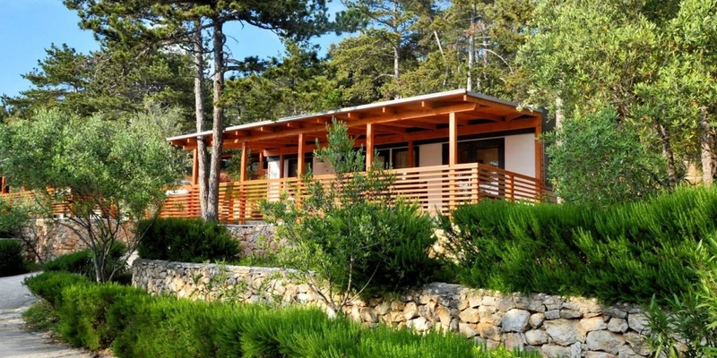 Wooden cabin with a large porch nestled among lush greenery and trees, surrounded by stone walls at Slatina