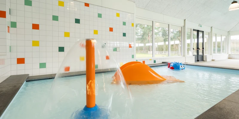 Water fountain spraying in a shallow indoor pool, surrounded by colorful walls and playful equipment at Vakantiepark de Veluwse Hoevegaerde