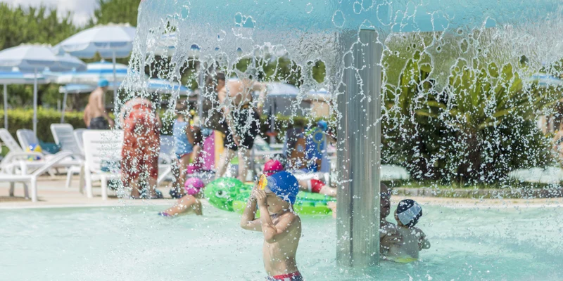 Children playing under a mushroom-shaped water fountain in a busy swimming pool area with deck chairs and umbrellas at Riva Nuova