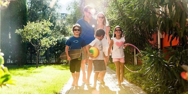 Family walks barefoot on a sunny path, surrounded by green trees and bushes, carrying toys at Holiday Park La Vela