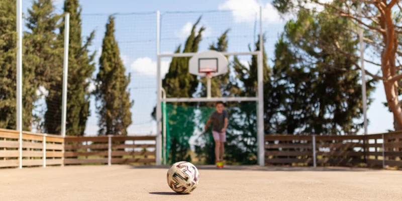 A soccer ball rests on a concrete court with a child running toward a basketball hoop in the background at Les Flamants Roses