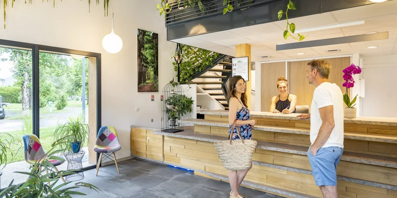 Three people converse at a bright reception desk in a plant-decorated room with large windows at La Nublière