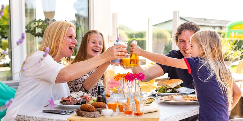 Family toasting drinks at an outdoor dining table, enjoying various dishes and flowers in the background at Europarcs de Woudhoeve