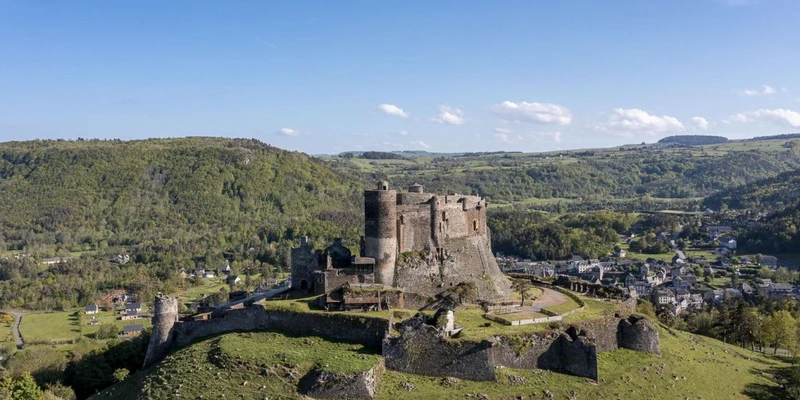 Ancient stone castle overlooks green valley, surrounded by scattered houses and distant wooded hills at La Ribeyre