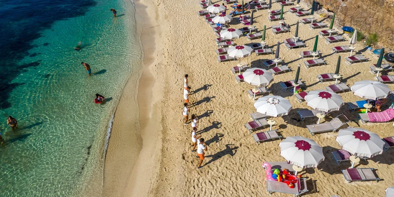 Beach umbrellas shading sun loungers scattered on sandy shore with people swimming in the clear blue ocean at Village Baia del Monaco
