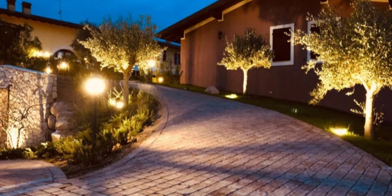 A paved pathway curves uphill, illuminated by garden lights and bordered by trees, leading to houses at La Bertoletta Village apartments