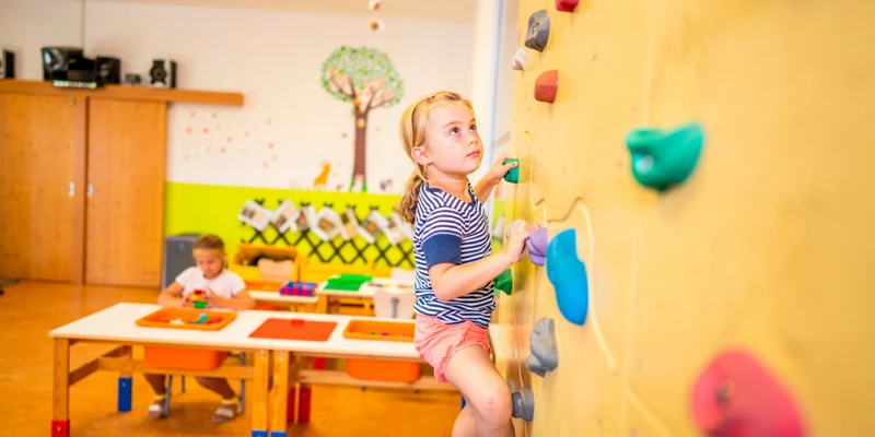 A child climbs an indoor wall while another child plays at a table, in a colorful classroom at Landal Brandnertal