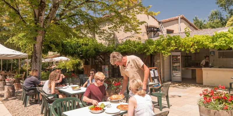 People dining outdoors while a waiter serves food; surrounded by greenery and rustic buildings at Le Camp de Florence