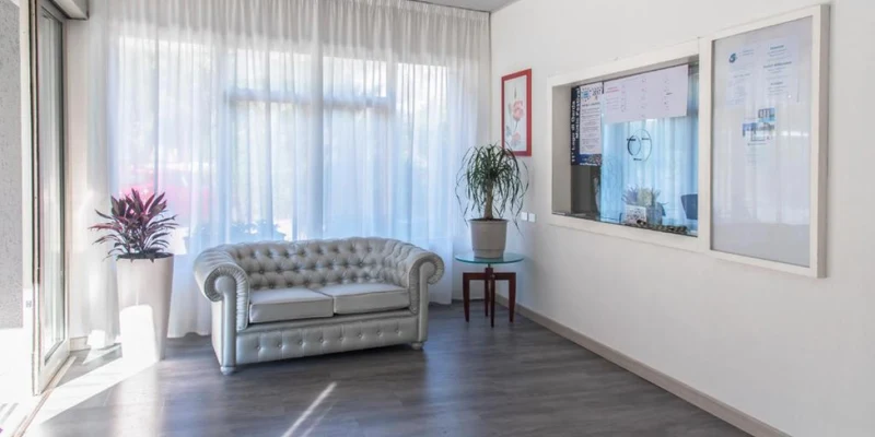 A grey couch sits next to potted plants in a bright, sunlit room with white curtains and framed artwork at Residence Monica