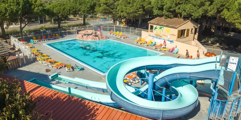A winding blue water slide leads into a large, sunlit swimming pool surrounded by colorful loungers and trees at La Maïre