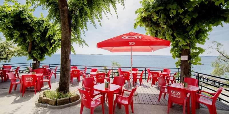 Red plastic chairs and tables sit under a red umbrella on a lakeside deck surrounded by trees at Camping Sereno
