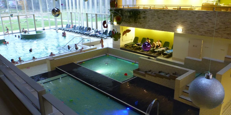 Indoor swimming pool with people relaxing in water, surrounded by lounge chairs, under a high ceiling with hanging decorations at Ferienresort Bad Bentheim