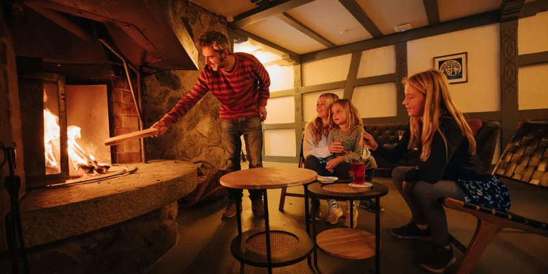 Man stoking a fireplace; family of three sitting on a couch with drinks, plate on a table; cozy living room at Jungfrau