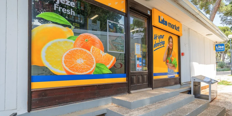 Grocery store facade displaying vibrant fruit imagery and promotional signage in a leafy outdoor setting at Porton Nature Hideouts