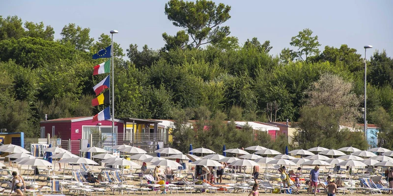Beachgoers relaxing on lounge chairs under umbrellas, near colorful cabins with flags, surrounded by lush greenery at San Francesco 