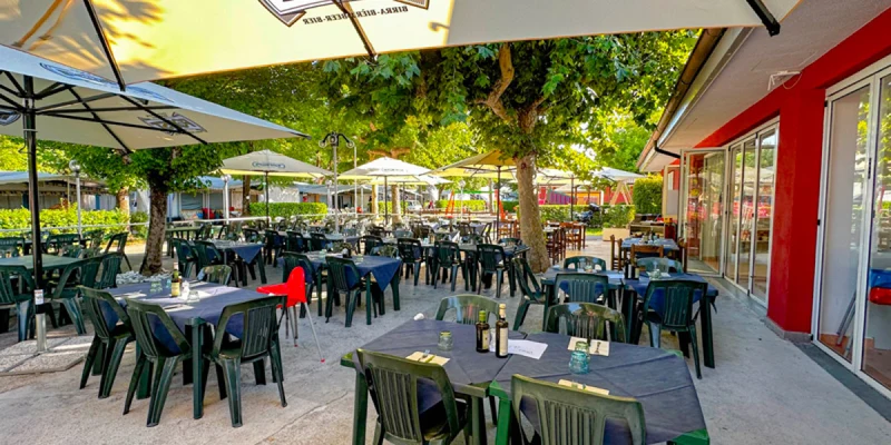Outdoor restaurant, tables with green chairs under large umbrellas beside red building, surrounded by trees and other dining areas at Camping Sereno