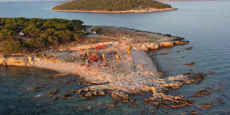 Colorful structures and people gathering on a rocky shoreline, with dense trees and a calm sea at Obonjan Island Resort