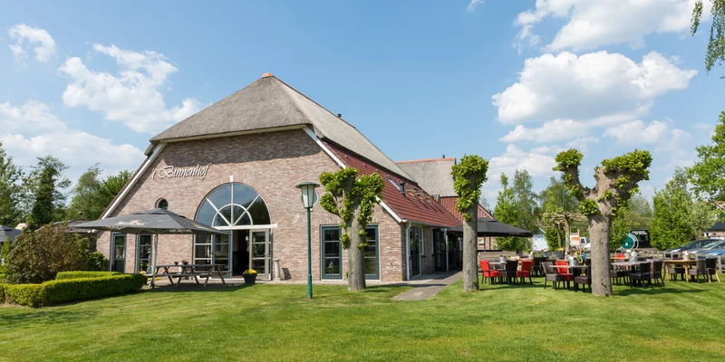 Brick building with arched windows, outdoor seating, and trimmed trees under a clear blue sky at Villapark de Weerribben