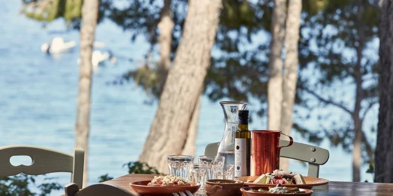 Plates of food and drinks on a wooden table surrounded by trees with a view of the sea at Candia Park Village