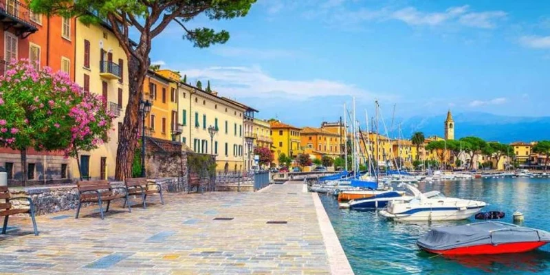 Boats docked along a waterfront promenade with colorful buildings and lush trees in the background at Domina Borgo Degli Ulivi