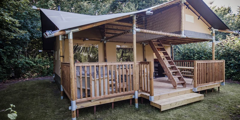 Wooden cabin with a shaded porch and upper loft, situated in a forested area at De Baalse Hei