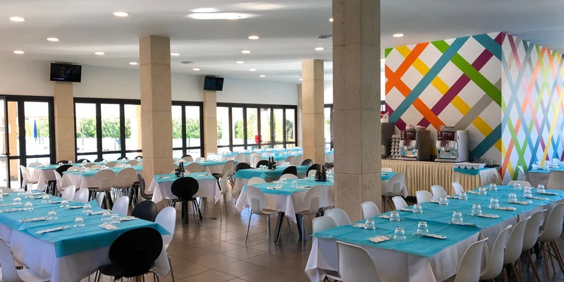 Tables set with turquoise cloths and white chairs in a bright dining hall; colorful geometric wall patterns at Belvedere Village