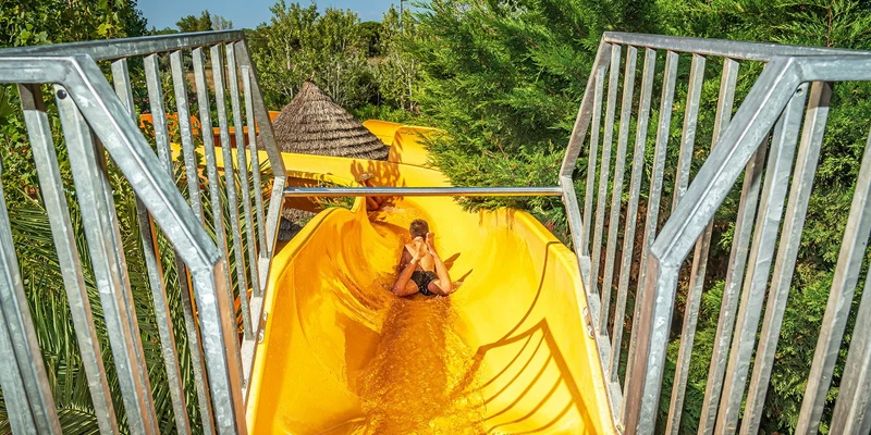 A child slides down a bright yellow water slide surrounded by lush green trees at Le Clos Virgile