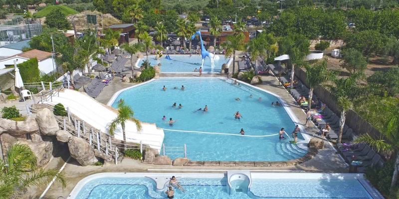 People swimming and relaxing in a large outdoor pool complex with slides, lounge chairs, and surrounded by trees at Alannia Els Prats