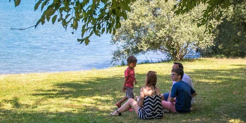Four people sitting and talking on grassy area near a lake with trees providing shade at Kanopée Camping & Lodge