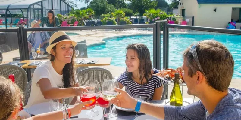Four people clinking glasses at a table by a pool surrounded by lush greenery and buildings at Marvilla Parks Pommeraie de l'Océan