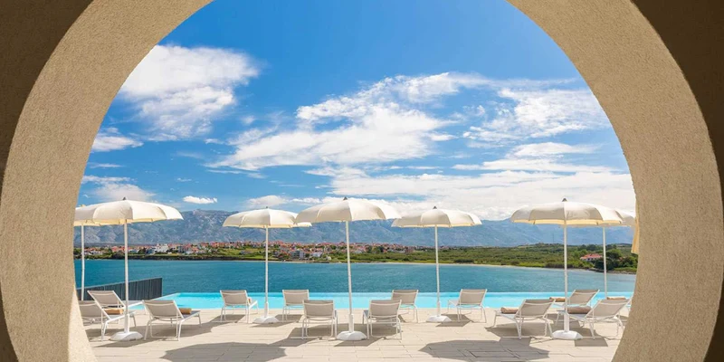 White umbrellas and lounge chairs are aligned by a pool, overlooking a scenic lake with mountains, viewed through an archway at Aminess Avalona Camping Resort