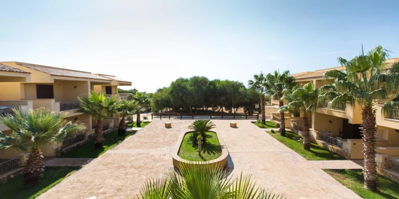 Buildings with balconies surround a pathway lined with palm trees and lush greenery at Triscina Mare