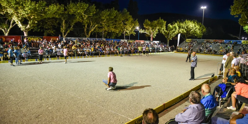 People watching a bocce game in an outdoor stadium with trees and night lights at Le Millau Plage