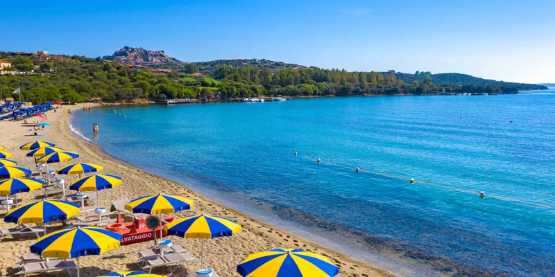Yellow-and-blue umbrellas line a sandy beach, overlooking a calm, clear blue sea, with distant green hills at Capo D'Orso