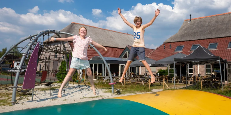 Two children jump joyfully on a large bouncing pillow, surrounded by a play structure and rustic buildings at Villapark de Weerribben
