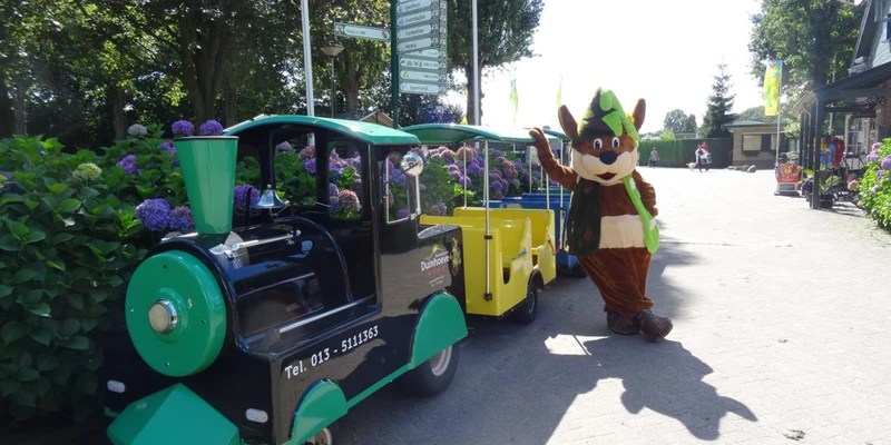 Mini-train with mascot waving near flowers and trees at Recreatiepark Duinhoeve