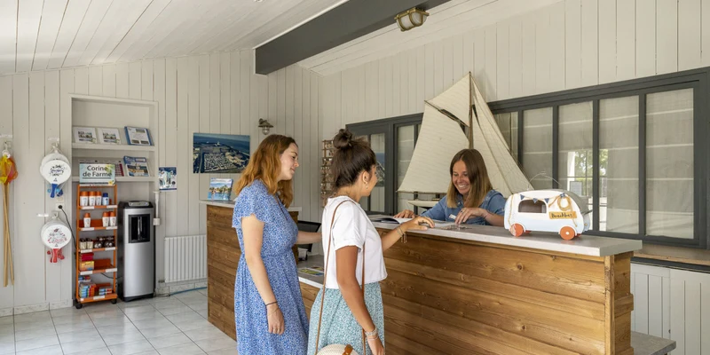 Two women inquire at a reception desk while an attendant assists them, inside a bright, nautical-themed room at La Plage